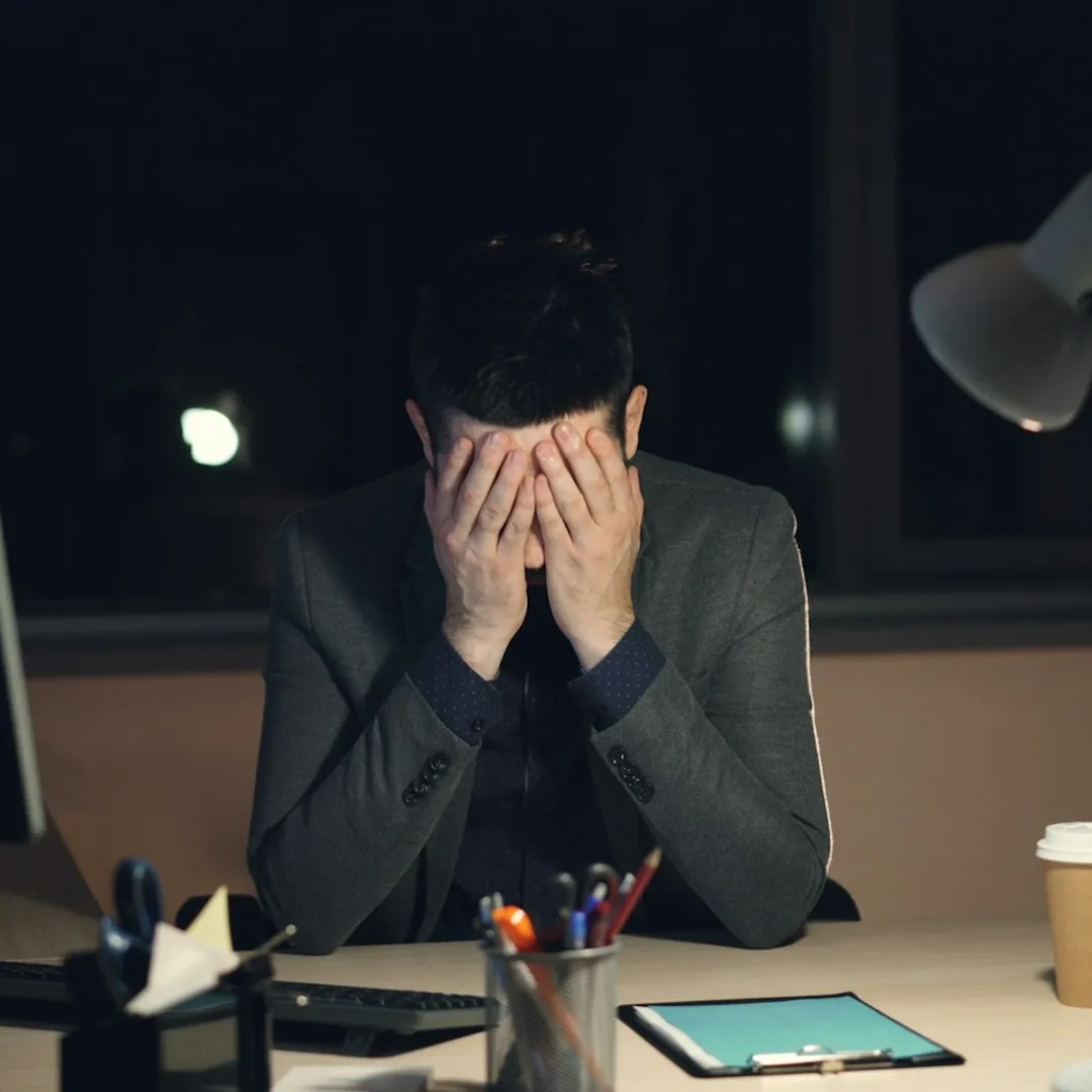 مردی با کت و شلوار پشت میز کار که شب دیروقت صورتش را میان دست‌هایش گرفته است